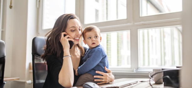 Career woman on phone at work while holding her baby who sits on her desk. 