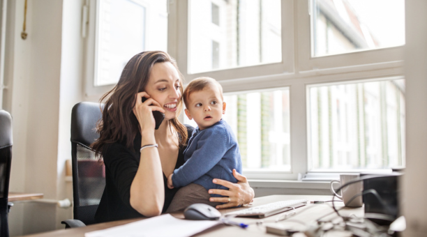 Career woman on phone at work while holding her baby who sits on her desk. 