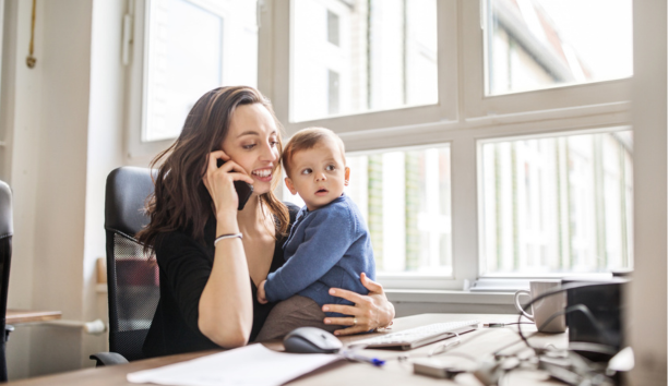 Career woman on phone at work while holding her baby who sits on her desk. 
