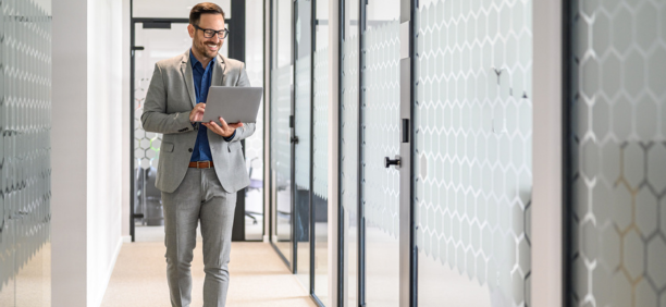 Professional man wearing glasses holding a laptop smiles and walks down hallway at work. 