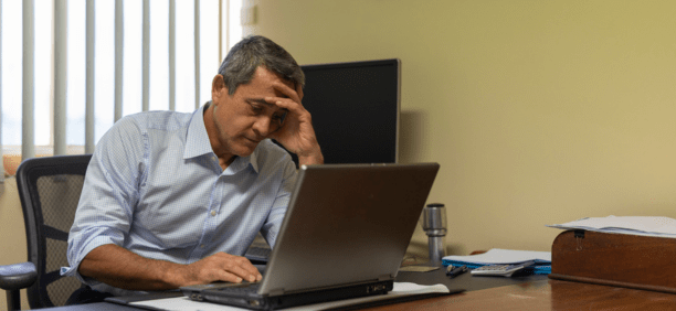 Minority man at work looking depressed while sitting at his desk on a laptop.