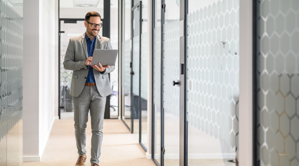 Professional man wearing glasses holding a laptop smiles and walks down hallway at work.
